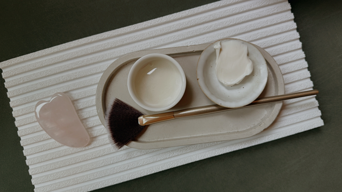 Jade roller, cream in small bowls, and a brush on a textured tray against a green background.